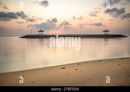 Sonnenaufgang über dem Meer, Blick vom Sandstrand bis zum Horizont, im Meer gibt es Wellenbrecher mit kleinen Tempeln, ruhiges Wasser mit kleinen Wellen und Reflexionen am tropischen Strand von Sanur, Bali, Indonesien Stockfoto