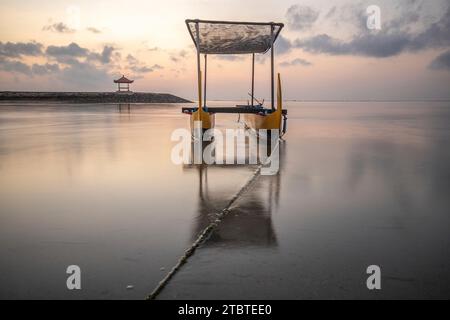 Morgenlandschaft auf einem Sandstrand, Blick über das Meer bis zum Horizont mit kleinen Tempeln im Wasser und einem Jukung, ein traditionelles Fischerboot im Meer, Sonnenaufgang in Sanur, Bali, Indonesien Stockfoto