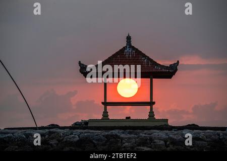Sonnenaufgang am Sandstrand mit kleinen Tempeln im Wasser, Landschaft mit roter kreisförmiger Sonne am Strand von Sanur, Bali, Indonesien Stockfoto