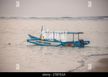 Morgenlandschaft auf einem Sandstrand, Blick über das Meer bis zum Horizont mit kleinen Tempeln im Wasser und einem Jukung, ein traditionelles Fischerboot im Meer, Sonnenaufgang in Sanur, Bali, Indonesien Stockfoto