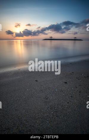 Sonnenaufgang über dem Meer, Blick vom Sandstrand bis zum Horizont, im Meer gibt es Wellenbrecher mit kleinen Tempeln, ruhiges Wasser mit kleinen Wellen und Reflexionen am tropischen Strand von Sanur, Bali, Indonesien Stockfoto