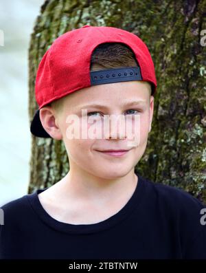 Kleiner Junge lehnt sich draußen an einen Baum. Er trägt seine Mütze nach hinten und schaut auf die Kamera. Er hat ein kleines Lächeln im Gesicht. Sommersprossen sprießen Stockfoto