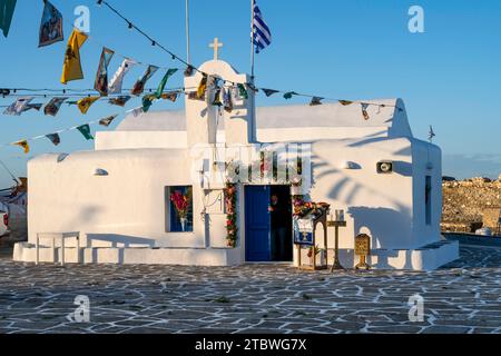 Weiße Kykladische griechisch-orthodoxe Kirche Agios Nikolaos bei Sonnenuntergang, dekoriert mit Fahnen, Hafen von Naoussa, Naoussa, Paros, Kykladen, Griechenland Stockfoto