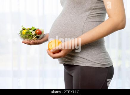 Stehende schwangere Frau, die Salat und Obst vor Vorhängen hält Stockfoto