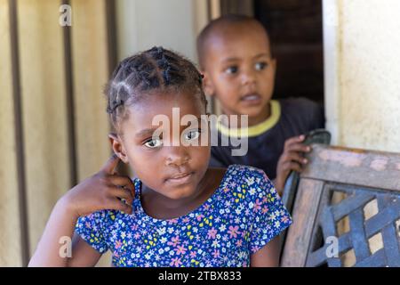 afrikanisches Mädchen mit Zöpfen und ihr Bruder spielt auf einer schmiedeeisernen Bank vor dem Haus Stockfoto