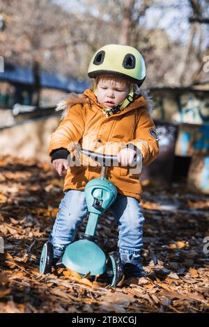 Entzückender Kleinkind mit gelbem Schutzhelm, der am Herbsttag im Freien auf dem Roller reitet. Kindertraining auf dem Minifahrrad im Stadtpark. Lustige Herbstaktivitäten im Freien für kleine Kinder Stockfoto