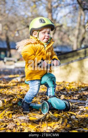 Entzückender Kleinkind mit gelbem Schutzhelm, der am Herbsttag im Freien auf dem Roller reitet. Kindertraining auf dem Minifahrrad im Stadtpark. Lustige Herbstaktivitäten im Freien für kleine Kinder Stockfoto