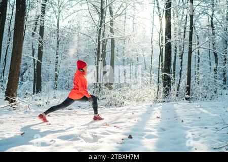 Die Trailläuferin trägt eine leuchtend rote, winddichte Jacke, die an einem sonnigen, frostigen Tag Dehnungsübungen im malerischen, verschneiten Wald macht. Sportlich aktiv Stockfoto