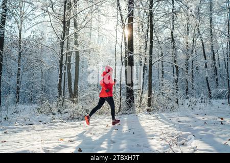 Trailläuferin in leuchtend roter, winddichter Jacke, die an einem sonnigen, frostigen Tag einen malerischen, verschneiten Waldweg joggen lässt. Sportlich aktive Menschen und Winter Stockfoto