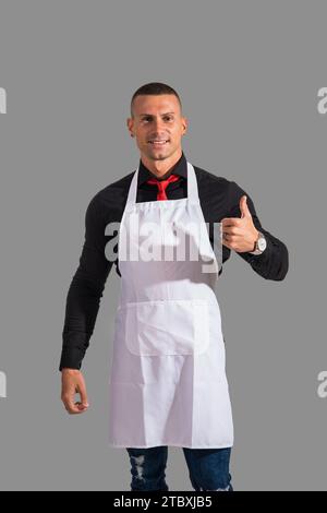 Attraktiver Koch oder Kellner posiert in weißer Schürze und schwarzem Hemd auf grauem Hintergrund, in Studioaufnahme Stockfoto