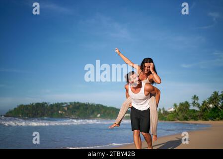 Fröhliches Paar genießt einen Huckepack-Ritt am sonnigen Strand. Der Mann trägt eine Frau auf dem Rücken, beide lächeln. Meereswellen, klarer Himmel, legere Sommerkleidung. Urlaub Stockfoto