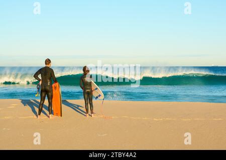 Ein Mann mit einem Bodyboard und ein Junge mit einem Surfbrett beobachten eine Welle, die am frühen Morgen in Trigg Beach, Perth, Western Australia, bricht. Stockfoto