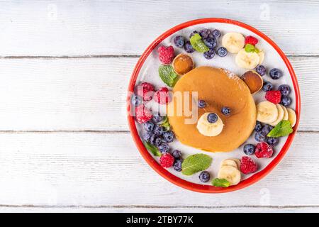 Lustige und süße Weihnachts-Teddybären-Pfannkuchen auf einem rot-weißen Teller, dekoriert mit frischen Beeren, Obst und Puderzucker, Kinderweihnachtsfrühstück Stockfoto