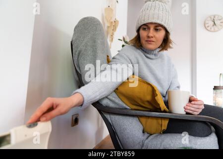 Eine Frau in warmen Kleidern hält ihre Hand auf dem Heizungsthermostat. Niedrige Heiztemperatur im Haus. Energiekrise-Konzept. Stockfoto