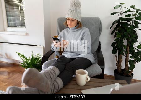 Eine Frau in warmer Kleidung mit Smartphone sitzt in der Nähe eines Heizstrahlers. Niedrige Temperatur im Haus. Energiekrise-Konzept. Stockfoto