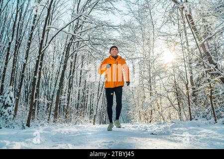 Lächelnder Trailläufer mittlerer Alters, gekleidet in leuchtend orangefarbener winddichter Jacke, hält an einem sonnigen, frostigen Tag schnell durch malerische, verschneite Wälder. S Stockfoto