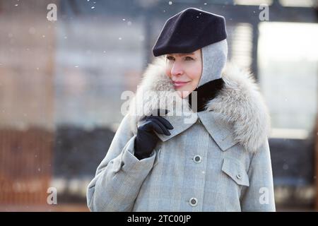 Porträt trendige reife Frau steht draußen im Schnee, trägt modisches mehrlagiges Winteroutfit, grauen Mantel mit Pelzkragen, Kapuze und stilvolle schwarze Kappe Stockfoto