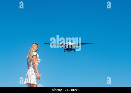 Coastal Encounter: Mädchen in weißem Kleid auf Steinen, Flugzeug in blauem Himmel Stockfoto