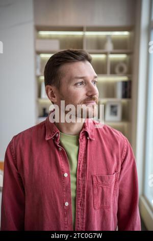 Nachdenklicher ruhiger positiver Mann, der steht, ins Fenster blickt, von Familie träumt, erfolgreiche Karriere Stockfoto