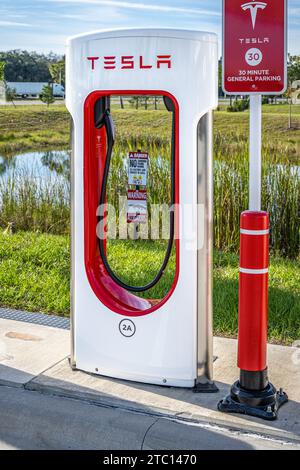 Tesla Supercharger EV-Station entlang der I-95 bei Buc-ees in Datona Beach, Florida, mit Alligator-Warnschild und interstate-Verkehr im Hintergrund. (USA) Stockfoto