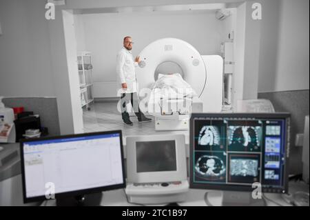 Medizinische Computertomographie oder MRT-Scanner. Arzt macht MRT, Patient liegt auf Liege. Männlicher Spezialist mit weißem Gewand und Brille, Blick in die Kamera, lächelnd. Konzept der modernen Diagnostik. Stockfoto