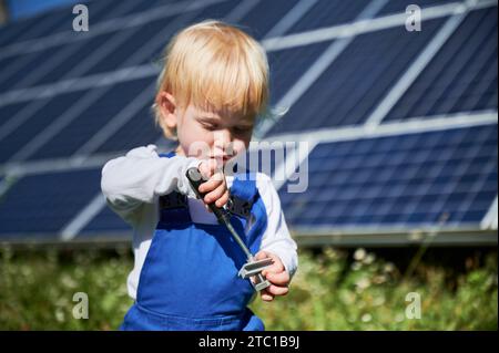 Kleines Kind, das den Schraubendreher auf dem Hintergrund von Solarpaneelen lernt. Neugieriger Junge, der an Reparaturen interessiert ist. Blondes Kleinkind versucht, die Schraube festzuziehen. Stockfoto