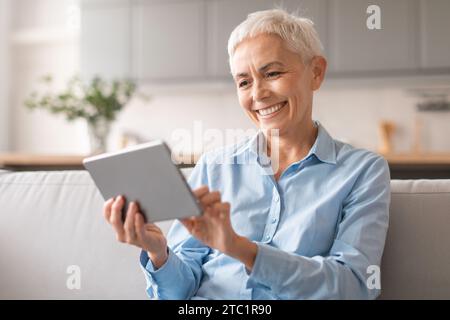 Lächelnde europäische reife Dame, die ein digitales Tablet hält und sich zu Hause entspannt Stockfoto