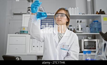 Schöne blonde Wissenschaftlerin in Aktion, junge Frau mit Schutzhandschuhen, die das Reagenzglas im High-Tech-Labor sorgfältig untersucht Stockfoto