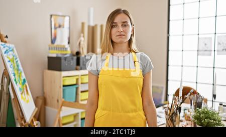 Tief in der Kreativität, schöne junge kaukasische Künstlerin, blond, intensiv in ihr Malhobby vertieft, stehend in ihrem Kunststudio, Pinsel und Stockfoto