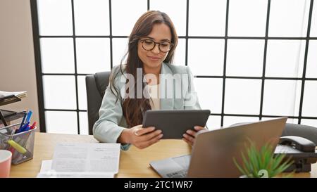 Selbstbewusste junge hispanische Frau, die im Büro erfolgreich arbeitet, schöne und glückliche Geschäftsangestellte mit Laptop und Touchpad am Schreibtisch Stockfoto