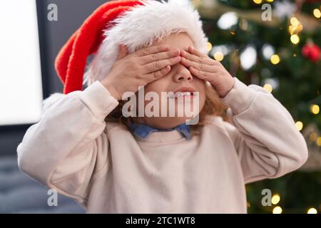 Entzückendes blondes Kleinkind, das die Augen bedeckt, mit Händen, die zu Hause am weihnachtsbaum stehen Stockfoto