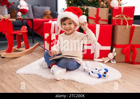 Ein entzückender hispanischer Junge, der den weihnachtsmann-Brief schreibt, sitzt zu Hause am weihnachtsbaum auf dem Boden Stockfoto