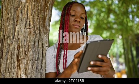 Coole afroamerikanische Frau, die sich auf ihr Touchpad fokussiert, sich im Stadtpark auf einen Baum lehnt, lässige Lifestyle-Atmosphäre im Grünen Stockfoto