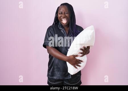 Junge afrikanische Frau, die Pijama trägt, das Kissen umarmt, die Zunge ausstreckt, glücklich mit lustigem Ausdruck. Emotionskonzept. Stockfoto