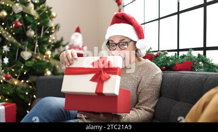 Frau mittleren Alters mit grauen Haaren, die weihnachtsmütze trägt, Geschenk mit überraschtem Gesicht zu Hause auspacken Stockfoto