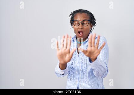 Afrikanische Frau mit Dreadlocks, die über weißem Hintergrund stehen und eine Brille tragen, die Angst und Angst hat, Ausdruck der Angst, stoppt die Geste mit den Händen, Shouti Stockfoto