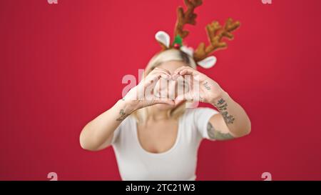 Junge schöne hispanische Frau mit Rentier-weihnachtshut, die Herzgeste über isoliertem roten Hintergrund macht Stockfoto