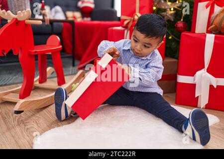 Entzückendes hispanisches Kleinkind, das zu Hause am weihnachtsbaum sitzt und ein Geschenk auspackt Stockfoto