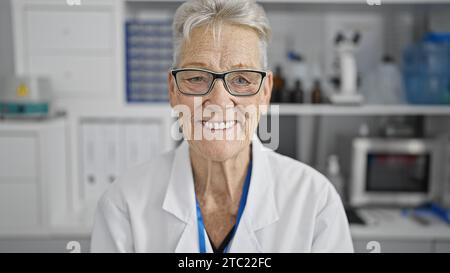 Selbstbewusste und lächelnde grauhaarige Wissenschaftlerin genießt ihre Arbeit und sitzt glücklich im Labor zwischen Mikroskop und Reagenzgläsern Stockfoto