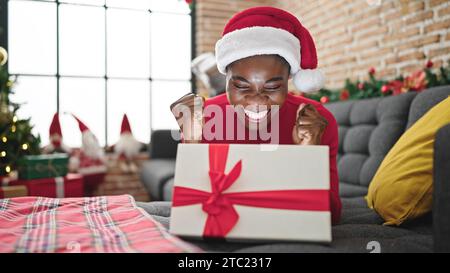 afroamerikanerin trägt weihnachtsmütze und packt ein Geschenk mit fröhlichem Gesicht zu Hause aus Stockfoto