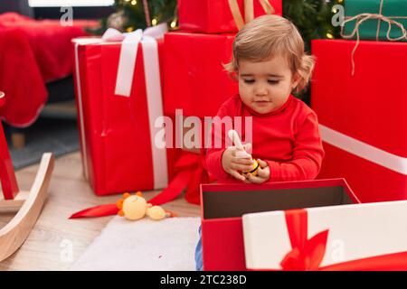 Entzückendes hispanisches Kleinkind, das zu Hause am weihnachtsbaum sitzt und ein Geschenk auspackt Stockfoto