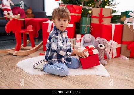 Entzückender hispanischer Junge, der zu Hause am weihnachtsbaum sitzt und ein Geschenk auspackt Stockfoto