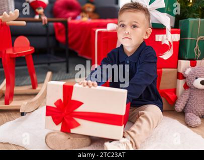 Entzückender hispanischer Junge, der zu Hause am weihnachtsbaum sitzt und ein Geschenk auspackt Stockfoto