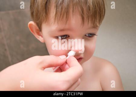Mit Glück reinigt die Frau ihr Gesicht und konzentriert sich auf die Nase mit einem Wattepad im Badezimmer. Kind im Alter von zwei Jahren (zweijähriger Junge) Stockfoto