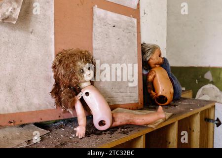Das Bild zeigt eine Puppe mit einer braunen Perücke und einem weißen Körper, die ihre Beine vermisst und auf einem Holzregal in einem Raum sitzt. Gruselige Spielzeuge in einem verlassenen Kindergarten Stockfoto