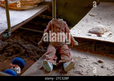 Eine kopflose Puppe mit roten Overalls, die auf einer staubigen Oberfläche sitzt. Kinderpuppe ohne Kopf. Ein dreckiges, kaputtes Kinderspielzeug von einer Strahlenkontamination Stockfoto