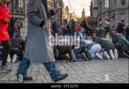 London / Großbritannien - 9. Dezember 2023: Ein Mann läuft an einer Gruppe muslimischer Männer vorbei, die auf dem Trafalgar Square nahe Big Ben bei einer pro-palästinensischen Demonstration beten Stockfoto