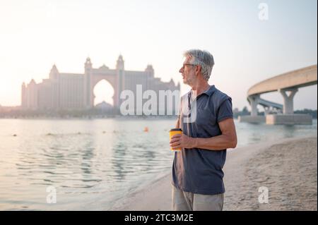 Senior kaukasischer Mann bei einer Tasse Kaffee und genießen die Schönheit von Palm Jumeirah in Dubai, gefangen vor der atemberaubenden Kulisse eines Sonnenuntergangs Stockfoto