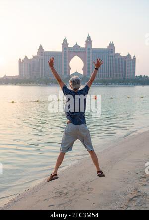 Senior kaukasischer Mann, der hoch in Palm Jumeirah in Dubai springt, gefangen vor der atemberaubenden Kulisse eines Sonnenuntergangs am öffentlichen Strand Stockfoto