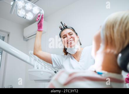 Zahnärztliche Klinikpatientententage auf der modernen medizinischen Station. Zahnarzt lächelt aufrichtig und zeigt Zahnlicht auf die junge Frau, bereit für die Zahnchirurgie Stockfoto